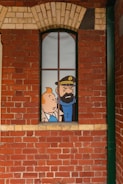 A comic-style illustration of two cartoon characters visible through a tall, narrow window set in a red brick wall. The character on the left is a young person with orange hair, and the one on the right is a bearded man wearing a hat with an anchor emblem.