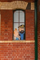 A comic-style illustration of two cartoon characters visible through a tall, narrow window set in a red brick wall. The character on the left is a young person with orange hair, and the one on the right is a bearded man wearing a hat with an anchor emblem.