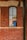 A comic-style illustration of two cartoon characters visible through a tall, narrow window set in a red brick wall. The character on the left is a young person with orange hair, and the one on the right is a bearded man wearing a hat with an anchor emblem.