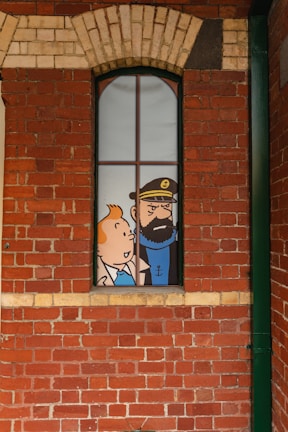 A comic-style illustration of two cartoon characters visible through a tall, narrow window set in a red brick wall. The character on the left is a young person with orange hair, and the one on the right is a bearded man wearing a hat with an anchor emblem.