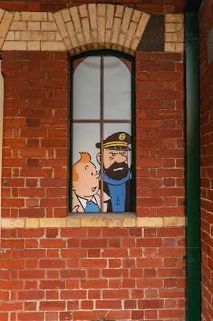 A comic-style illustration of two cartoon characters visible through a tall, narrow window set in a red brick wall. The character on the left is a young person with orange hair, and the one on the right is a bearded man wearing a hat with an anchor emblem.