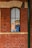 A comic-style illustration of two cartoon characters visible through a tall, narrow window set in a red brick wall. The character on the left is a young person with orange hair, and the one on the right is a bearded man wearing a hat with an anchor emblem.