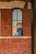 A comic-style illustration of two cartoon characters visible through a tall, narrow window set in a red brick wall. The character on the left is a young person with orange hair, and the one on the right is a bearded man wearing a hat with an anchor emblem.