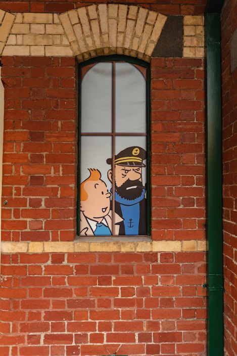 A comic-style illustration of two cartoon characters visible through a tall, narrow window set in a red brick wall. The character on the left is a young person with orange hair, and the one on the right is a bearded man wearing a hat with an anchor emblem.