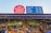 A bustling market scene with a variety of colorful fruits displayed in neatly arranged stalls. Shoppers browse through the produce under a covered structure adorned with decorative metalwork. A prominent red sign indicates the name, 'South Melbourne Market,' and a blue sign displays the opening hours for different days.