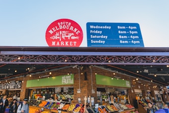 A bustling market scene with a variety of colorful fruits displayed in neatly arranged stalls. Shoppers browse through the produce under a covered structure adorned with decorative metalwork. A prominent red sign indicates the name, 'South Melbourne Market,' and a blue sign displays the opening hours for different days.