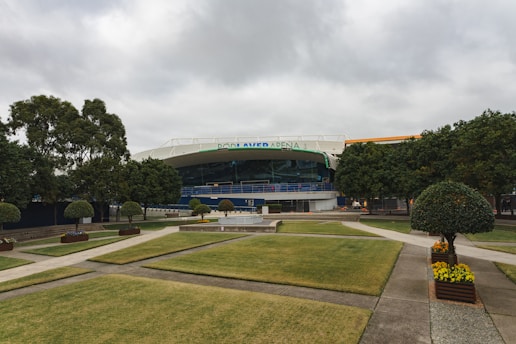 An outdoor scene featuring an arena in the background surrounded by neatly manicured grass lawns and several well-pruned trees. Paved walkways crisscross the area leading towards the arena, and planters with yellow flowers adorn the path. The sky is overcast, adding a muted tone to the ambiance.