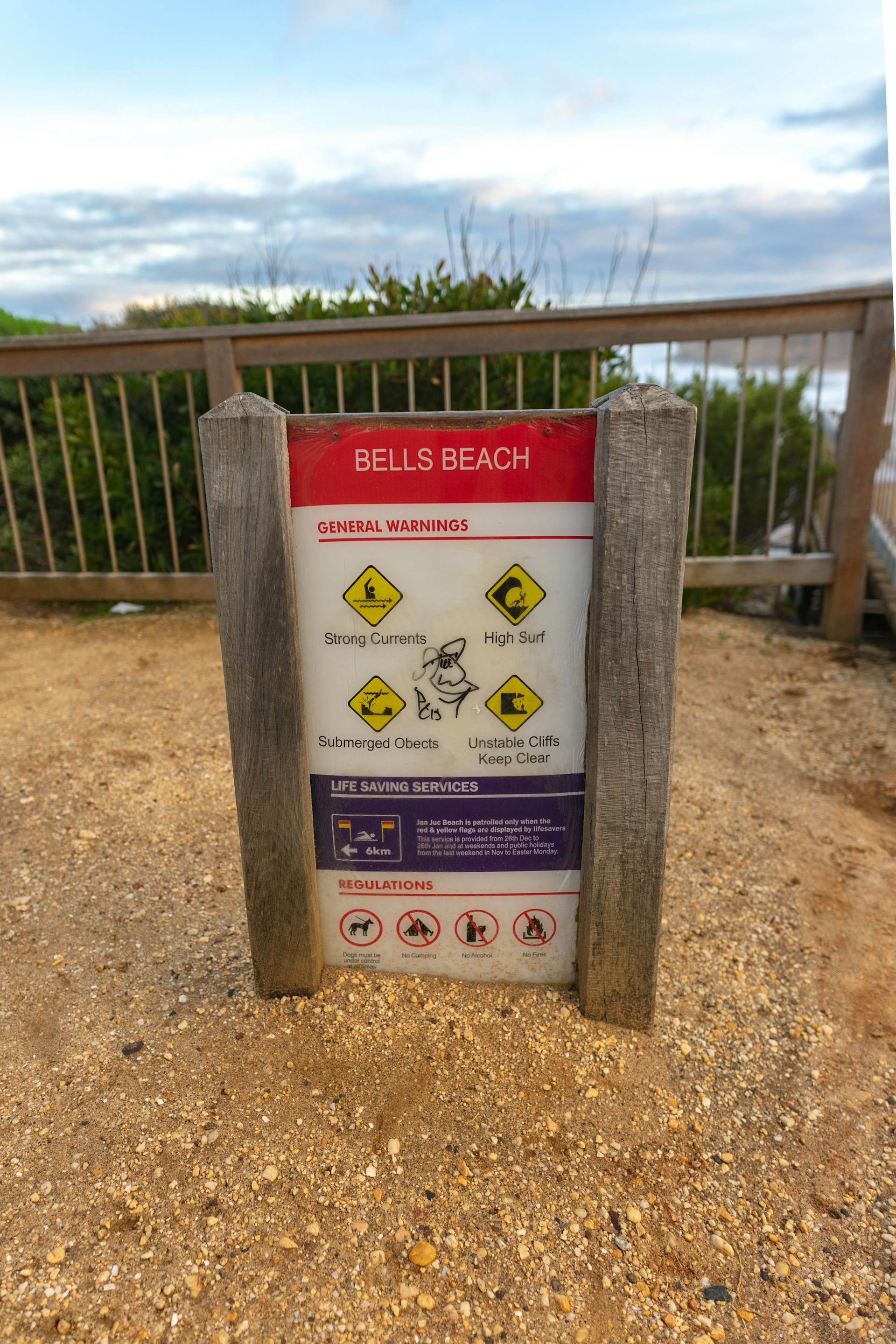 A beach warning sign situated on a sandy path with a wooden fence and vegetation in the background. The sign displays a series of warning symbols for strong currents, high surf, submerged objects, and unstable cliffs. It also contains safety information and regulations, indicating it’s a place with potentially hazardous conditions.