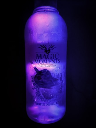 A bottle of Magic Moments pure grain vodka is depicted, illuminated with a glowing purple light. The bottle is frosty, suggesting it is cold, and features a design of a guitar with bubbles around it and the label reading 'Magic Moments Pure Grain Vodka' and 'Triple Distilled'.
