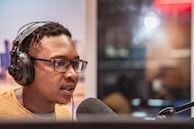 A person wearing headphones and glasses is speaking into a microphone, possibly in a recording studio. The background is blurred, suggesting a professional or indoor setting.