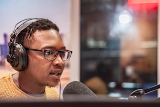 A person wearing headphones and glasses is speaking into a microphone, possibly in a recording studio. The background is blurred, suggesting a professional or indoor setting.