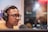 A person wearing headphones and glasses is speaking into a microphone, possibly in a recording studio. The background is blurred, suggesting a professional or indoor setting.