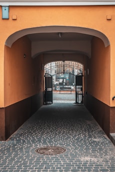 A narrow arched passageway made of orange and brown walls leads to an open area through an ornate metal gate. Cobblestone pavement covers the ground, and there is a distant view of a bench and outdoor furniture.