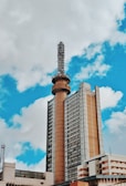 A tall communication tower rises from the center of a large, multi-story building with a modern architectural design. The sky is partly cloudy, featuring a mix of white clouds and blue sky.