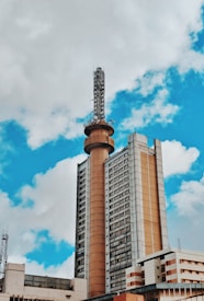 A tall communication tower rises from the center of a large, multi-story building with a modern architectural design. The sky is partly cloudy, featuring a mix of white clouds and blue sky.