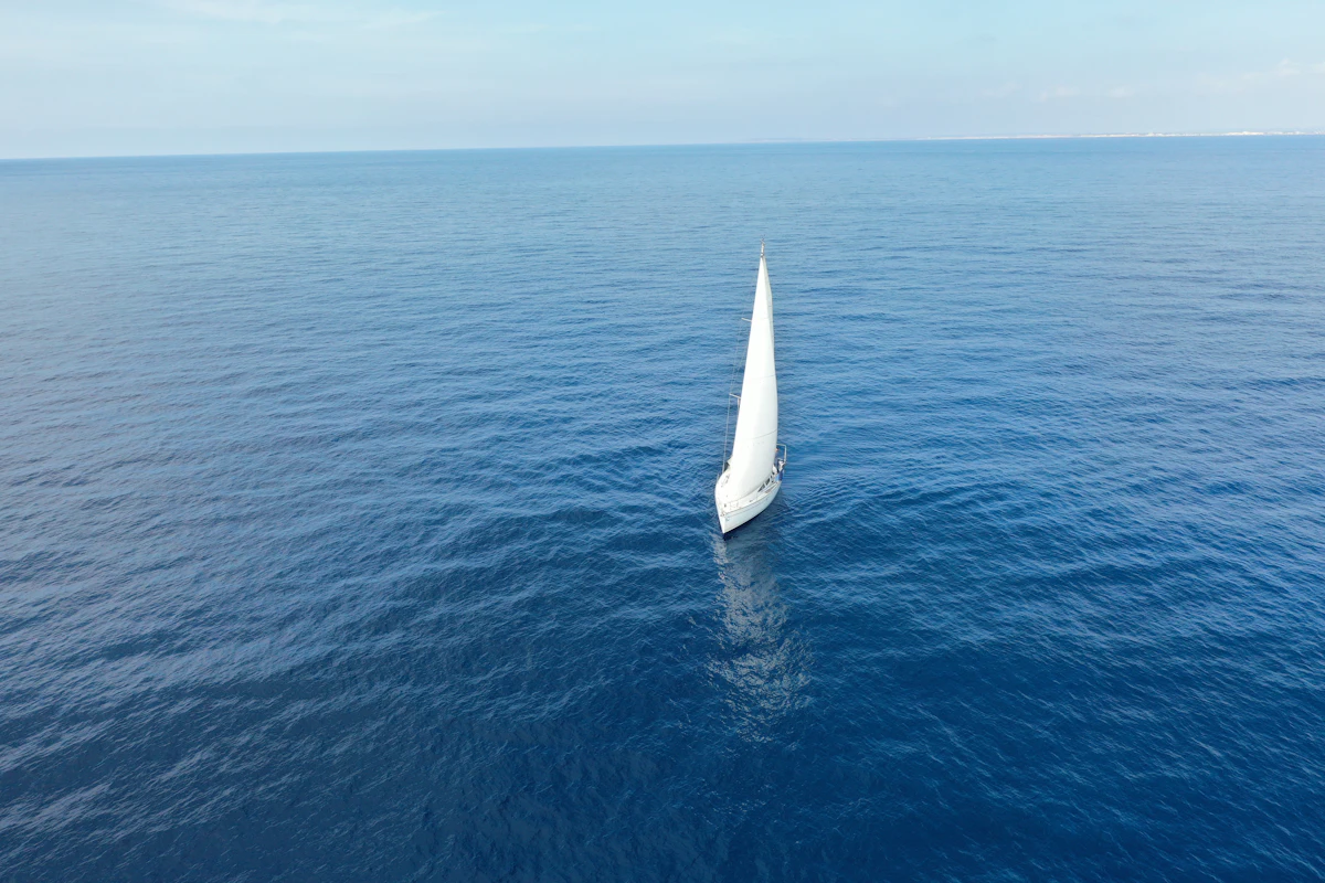 A single white sloop under sail alone on flat deep-blue open water, seen from high on the shore, no other boat in sight.