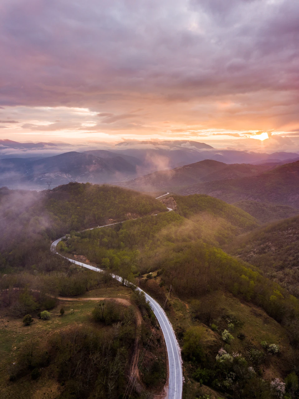 The image presents an aerial view of a winding road that cuts through a verdant landscape, surrounded by lush green hills and mountains. The sky above is painted with hues of orange and pink, suggesting either sunrise or sunset. The road curves gently to the right in the foreground, disappearing into the distance as it winds its way around the hillside.

The surrounding area is densely populated with trees and vegetation, adding a sense of tranquility and natural beauty to the scene. The image is taken from an aerial perspective, providing a comprehensive view of the winding road and the expansive landscape beyond.