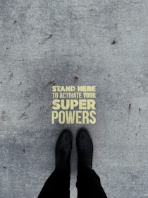 A pair of black boots standing on a concrete surface with a motivational message stenciled in yellow that reads 'Stand Here to Activate Your Super Powers'.