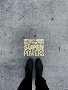 A pair of black boots standing on a concrete surface with a motivational message stenciled in yellow that reads 'Stand Here to Activate Your Super Powers'.