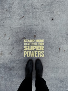 A pair of black boots standing on a concrete surface with a motivational message stenciled in yellow that reads 'Stand Here to Activate Your Super Powers'.