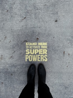 A pair of black boots standing on a concrete surface with a motivational message stenciled in yellow that reads 'Stand Here to Activate Your Super Powers'.