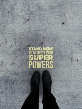 A pair of black boots standing on a concrete surface with a motivational message stenciled in yellow that reads 'Stand Here to Activate Your Super Powers'.