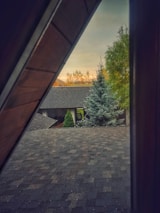 A slanted view through an attic window reveals rooftops and a serene landscape in the background. A lush evergreen tree and well-maintained bushes are visible close to a house with dark shingles. The evening sky casts a warm, golden hue over the scene, suggesting a sunset or sunrise.