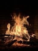 A warm and vibrant flame burning in a fireplace, surrounded by dark bricks. The logs beneath the fire are partially charred, with bright, flickering flames rising upward.