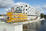 A modern waterfront urban scene featuring a large contemporary white building with numerous windows standing next to a construction site. Bright yellow temporary office units with red accents are set up near the water, displaying the logo of a construction company. Various construction vehicles and equipment are visible, along with graffiti along the water’s edge.