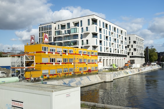 A modern waterfront urban scene featuring a large contemporary white building with numerous windows standing next to a construction site. Bright yellow temporary office units with red accents are set up near the water, displaying the logo of a construction company. Various construction vehicles and equipment are visible, along with graffiti along the water’s edge.