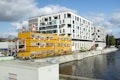A modern waterfront urban scene featuring a large contemporary white building with numerous windows standing next to a construction site. Bright yellow temporary office units with red accents are set up near the water, displaying the logo of a construction company. Various construction vehicles and equipment are visible, along with graffiti along the water’s edge.
