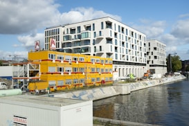 A modern waterfront urban scene featuring a large contemporary white building with numerous windows standing next to a construction site. Bright yellow temporary office units with red accents are set up near the water, displaying the logo of a construction company. Various construction vehicles and equipment are visible, along with graffiti along the water’s edge.