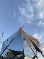 A modern, glass-covered building under construction with a clear blue sky in the background. Cranes and construction equipment are visible, working on the upper sections of the structure. The glass reflects the surroundings and the sky, adding a dynamic visual element.