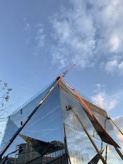 A modern, glass-covered building under construction with a clear blue sky in the background. Cranes and construction equipment are visible, working on the upper sections of the structure. The glass reflects the surroundings and the sky, adding a dynamic visual element.