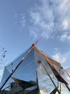 A modern, glass-covered building under construction with a clear blue sky in the background. Cranes and construction equipment are visible, working on the upper sections of the structure. The glass reflects the surroundings and the sky, adding a dynamic visual element.