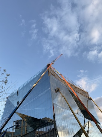 A modern, glass-covered building under construction with a clear blue sky in the background. Cranes and construction equipment are visible, working on the upper sections of the structure. The glass reflects the surroundings and the sky, adding a dynamic visual element.