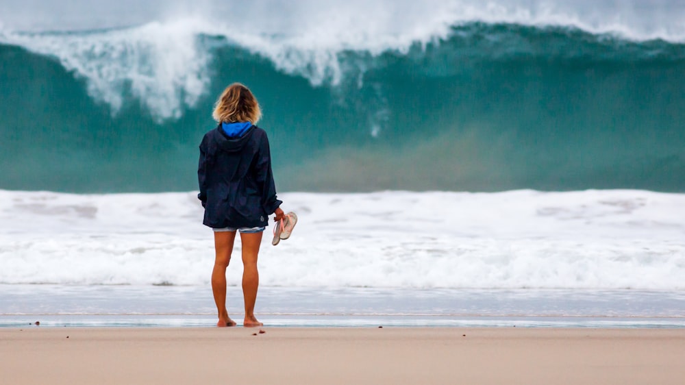 barefooted person holding flip-flops while standing near seashore during daytime