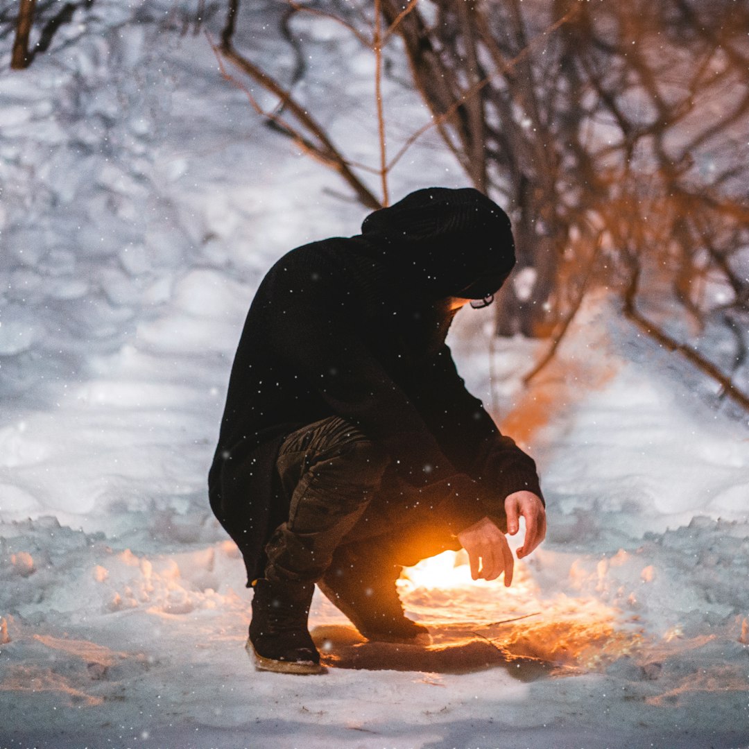 person wearing black jacket almost sitting on snowy field near bonfire during daytime