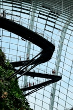 An indoor walkway with people walking along it is integrated into a lush, green vertical garden. The structure is enclosed within a large, glass-paneled dome, creating a modern and open atmosphere.