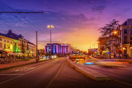 people walking on pathway near buildings and different vehicles on road during night time in Arnhem Netherlands