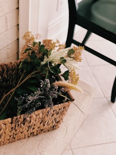 A woven rattan decorative basket filled with dried wildflowers on a rustic wooden floor.