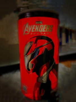 A vibrant red can features bold graphics and text related to Marvel's Avengers. A stylized helmet design is prominently displayed with sharp, angular lines.