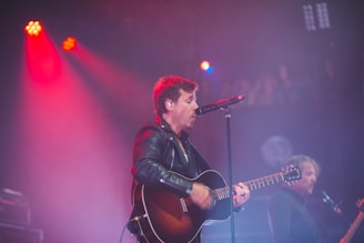A man is performing on stage, playing an acoustic guitar and singing into a microphone. He is wearing a leather jacket, and the stage is lit with red and purple lights. Another musician is visible in the background, also holding a guitar.