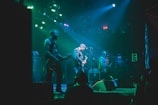 A band is performing on stage with three visible members playing electric guitars under vibrant green and blue stage lights. The atmosphere is energetic, with the audience barely visible in the darker areas, suggesting a lively concert.