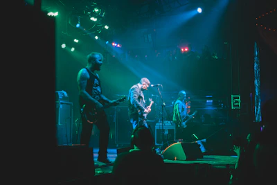 A band is performing on stage with three visible members playing electric guitars under vibrant green and blue stage lights. The atmosphere is energetic, with the audience barely visible in the darker areas, suggesting a lively concert.
