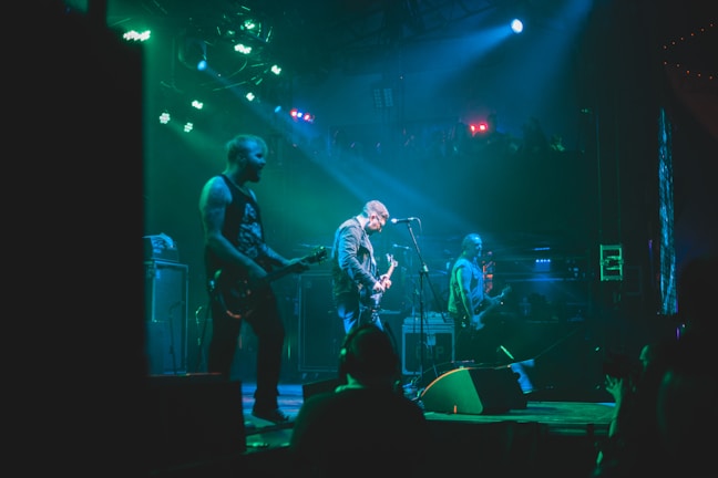 A band is performing on stage with three visible members playing electric guitars under vibrant green and blue stage lights. The atmosphere is energetic, with the audience barely visible in the darker areas, suggesting a lively concert.