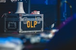 A large, rugged metal road case with reinforced corners and handles, marked with the yellow letters 'OLP' on its side. It rests on a stage floor, accompanied by a white funnel-like object and various cables. The background has a dimly lit, blurred environment, suggesting a performance or backstage setup.