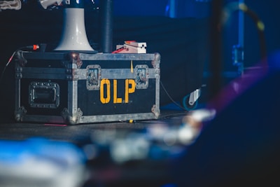 A large, rugged metal road case with reinforced corners and handles, marked with the yellow letters 'OLP' on its side. It rests on a stage floor, accompanied by a white funnel-like object and various cables. The background has a dimly lit, blurred environment, suggesting a performance or backstage setup.