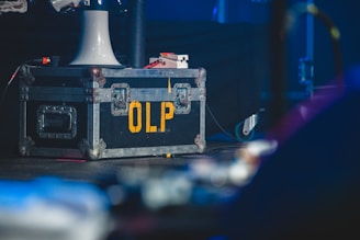 A large, rugged metal road case with reinforced corners and handles, marked with the yellow letters 'OLP' on its side. It rests on a stage floor, accompanied by a white funnel-like object and various cables. The background has a dimly lit, blurred environment, suggesting a performance or backstage setup.