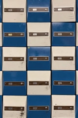 Rows of metal mailboxes with alternating blue and white doors, each labeled with a number and equipped with a lock. Some doors show marks and scratches.